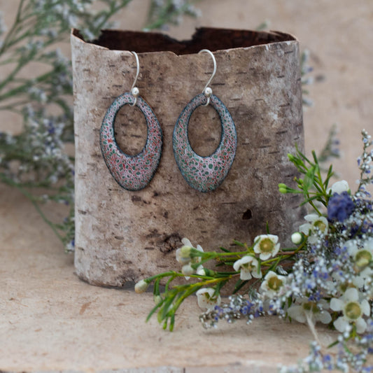 Handmade oval enamel earrings in soft green, pink, and lavender tones with a delicate lace-like pattern, displayed on a birch bark stand with wildflowers, by Divella Designs.