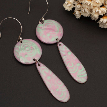 A pair of pink and green enamel copper earrings with a geometric pattern, displayed against a dark background with a decorative flower in the background.