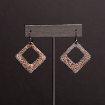 Square copper enamel earrings with soft sage green, rust, and ivory crackle glaze, displayed on an earring stand against a dark gray background.