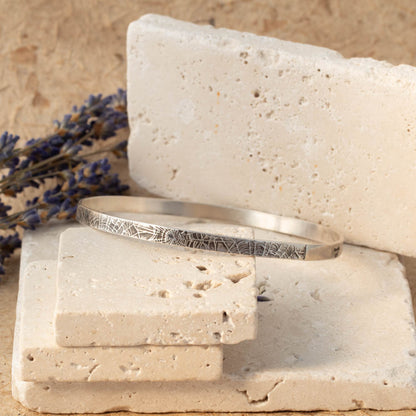 Handmade sterling silver spiderweb textured bangle resting on stacked stone blocks with soft neutral tones and lavender in the background.