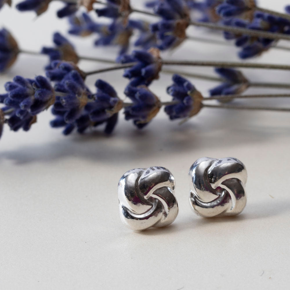 Polished sterling silver knot stud earrings displayed on a light surface with lavender flowers in the background.