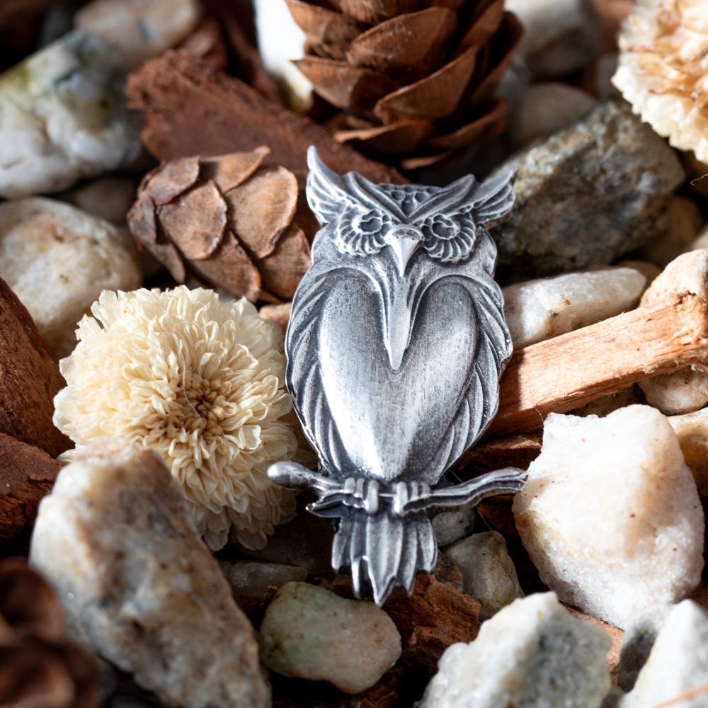 A sterling silver metal owl pendant or brooch with detailed feather and facial engravings, displayed among natural stones, pinecones, and dried flowers.