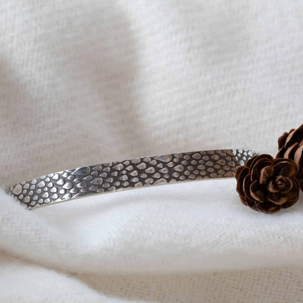 Sterling silver cuff bracelet with a raindrop texture, displayed on soft white fabric with small pinecones for decoration.