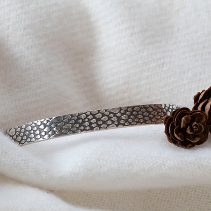 Sterling silver cuff bracelet with a raindrop texture, displayed on soft white fabric with small pinecones for decoration.