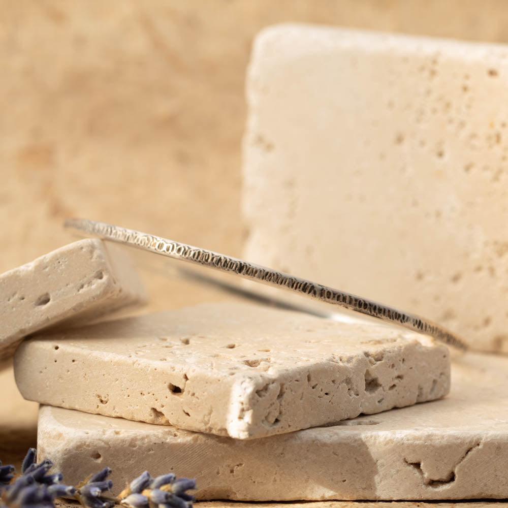 Handmade sterling silver textured bangle photographed on stacked stone blocks with soft neutral styling.