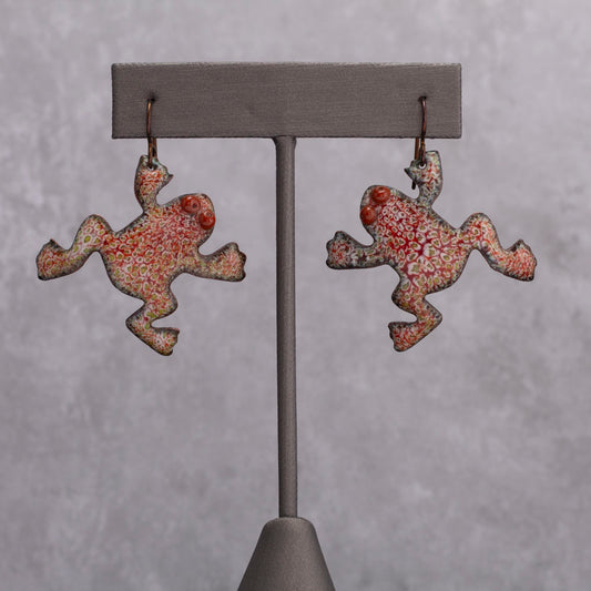 Playful red and green enamel frog-shaped earrings with dotted patterns and raised spiral eyes, displayed on a gray stand against a light gray background.