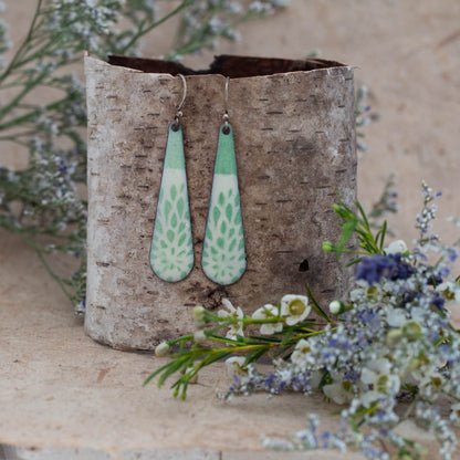 Handcrafted enamel earrings in a soft teardrop shape featuring mint green and white botanical thistle-inspired patterns. Lightweight dangles with a fresh spring feel, displayed on natural birch bark with wildflowers – Spring Thistle Earrings by Divella Designs.