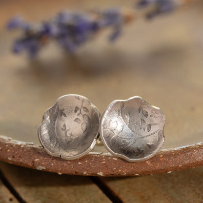 Silver floral earrings on a textured surface with blurred purple flowers in the background