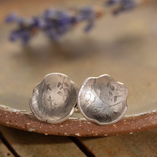Silver floral earrings on a textured surface with blurred purple flowers in the background