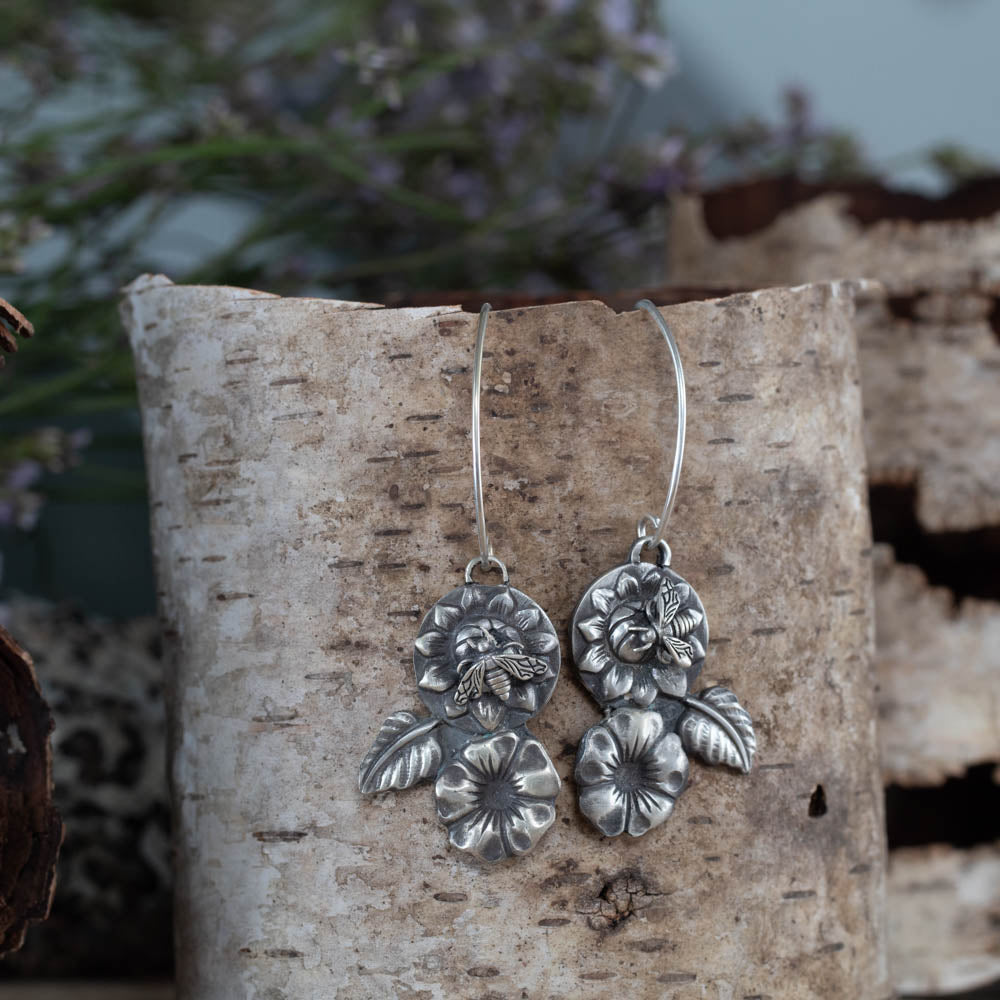 Sterling silver bee and flower dangle earrings by Divella Designs, 2" long with sculpted blooms, leaves, and patina finish, handmade in Vermont, shown with birch bark