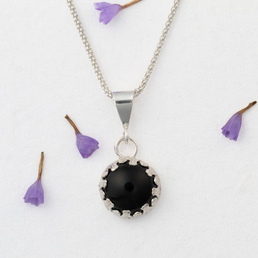 Sterling silver pendant necklace featuring a round black onyx stone in a scalloped bezel setting, displayed on a white background with scattered purple flower petals