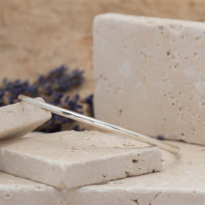 Handmade sterling silver faceted bangle with subtle hammered texture, photographed on stacked stone blocks with neutral tones and lavender in the background.