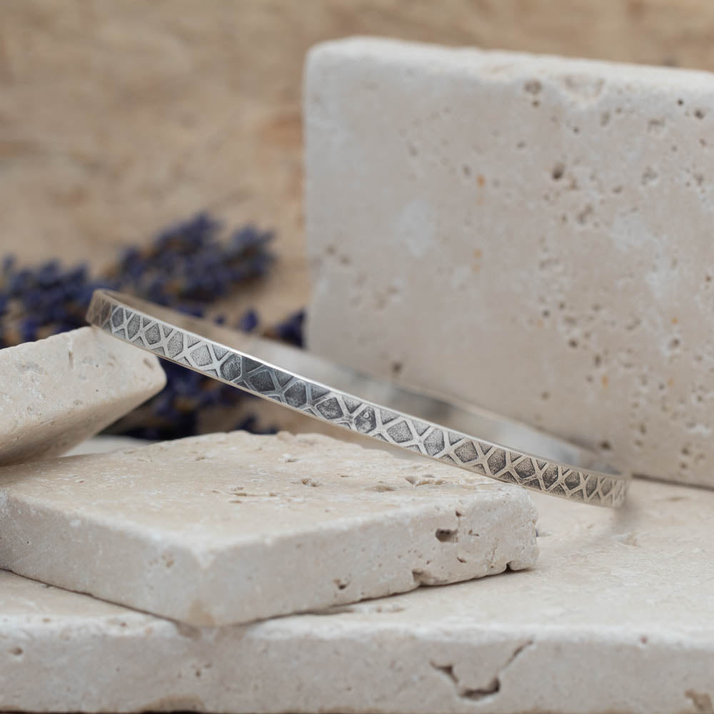 Handmade sterling silver geometric patterned bangle with oxidized texture, photographed on stacked stone blocks with neutral tones and lavender in the background.