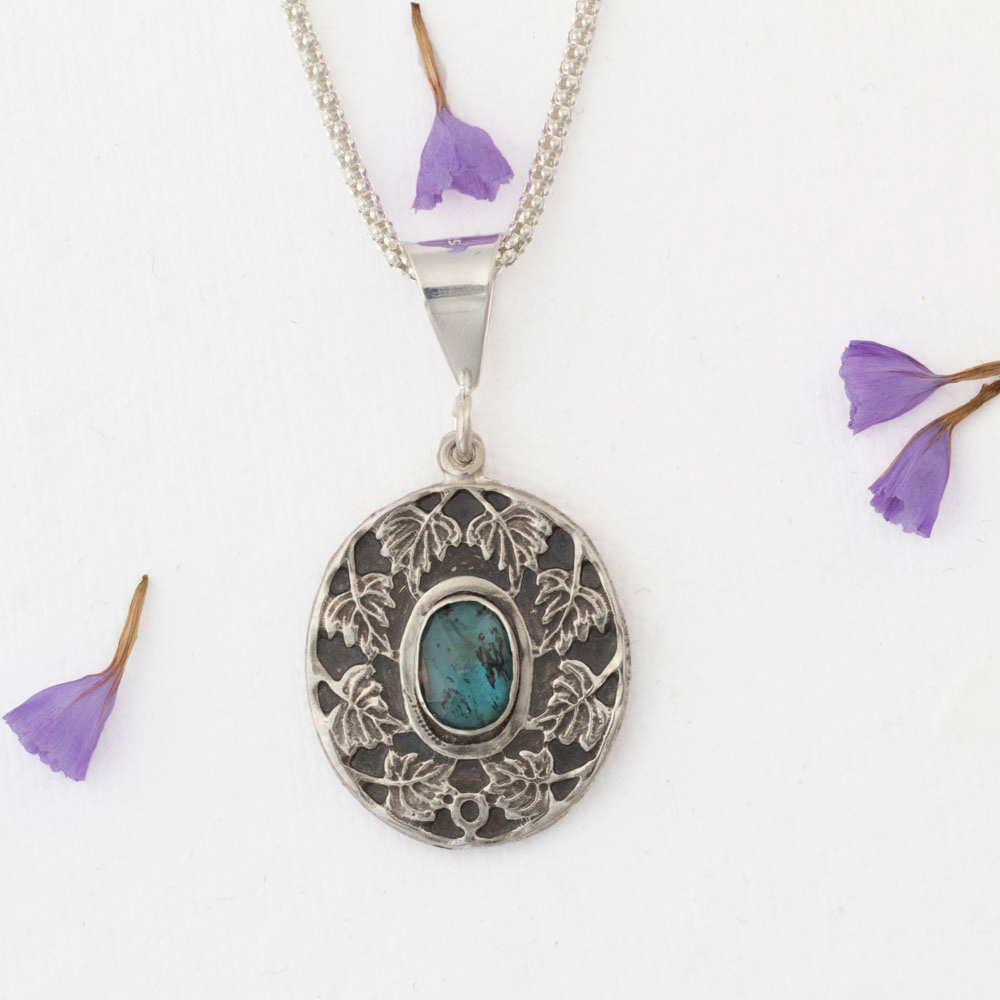 Sterling silver pendant necklace with an ornate leaf design surrounding an oval blue-green kyanite stone, shown on a white background with scattered purple flower petals.