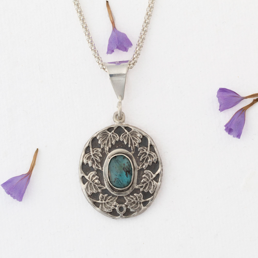 Sterling silver pendant necklace with an ornate leaf design surrounding an oval blue-green kyanite stone, shown on a white background with scattered purple flower petals.