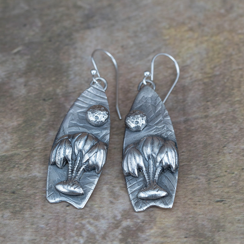 Handcrafted sterling silver earrings featuring sculpted willow trees under a textured full moon on leaf-embossed backdrops – Moonlight Surfing nature-inspired earrings by Divella Designs, shown on earthy background.