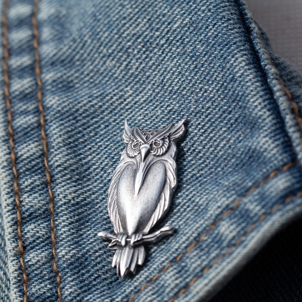 A silver owl-shaped brooch or pin displayed on a blue denim jacket, showing the detailed metalwork against the blue fabric background.