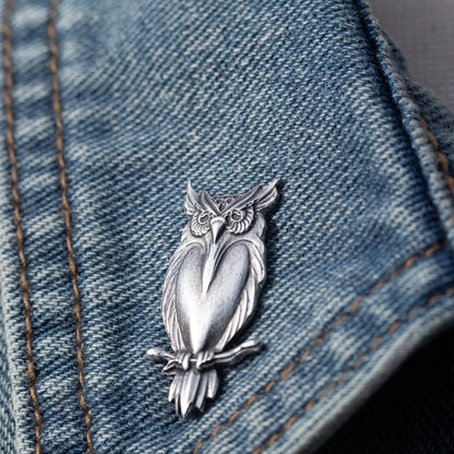 A silver owl-shaped brooch or pin displayed on a blue denim jacket, showing the detailed metalwork against the blue fabric background.