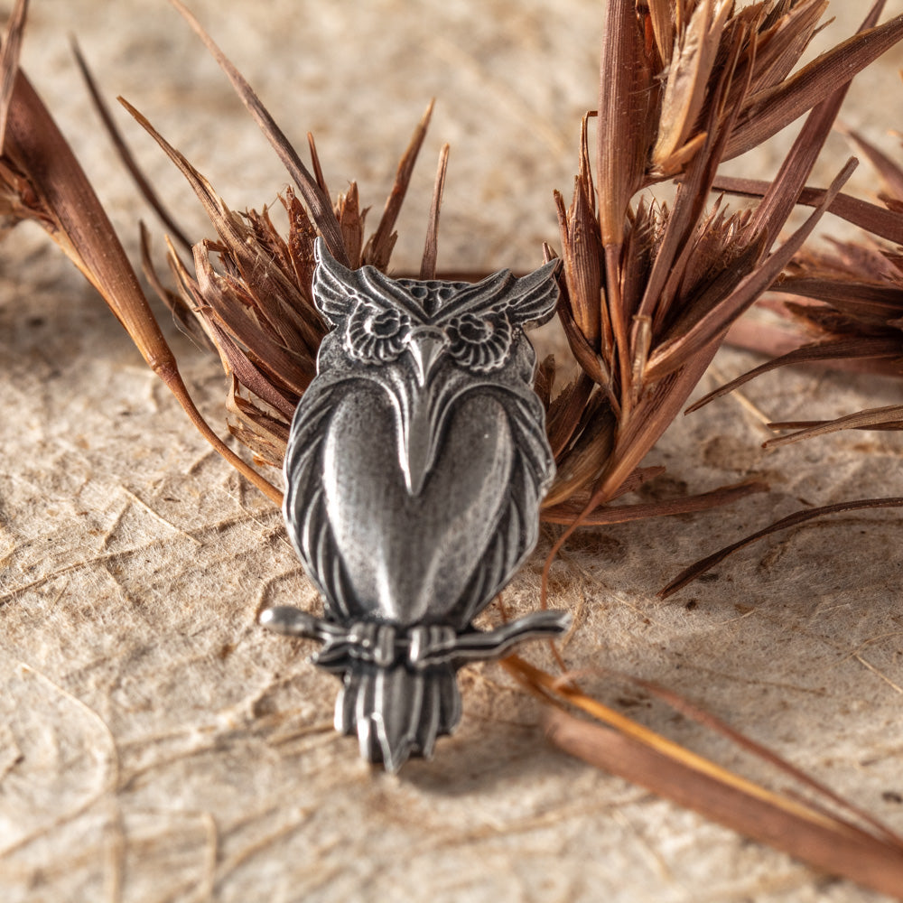 An antiqued silver owl pendant or charm perched on a branch, displayed on a textured beige surface with dried brown plant stems behind it.