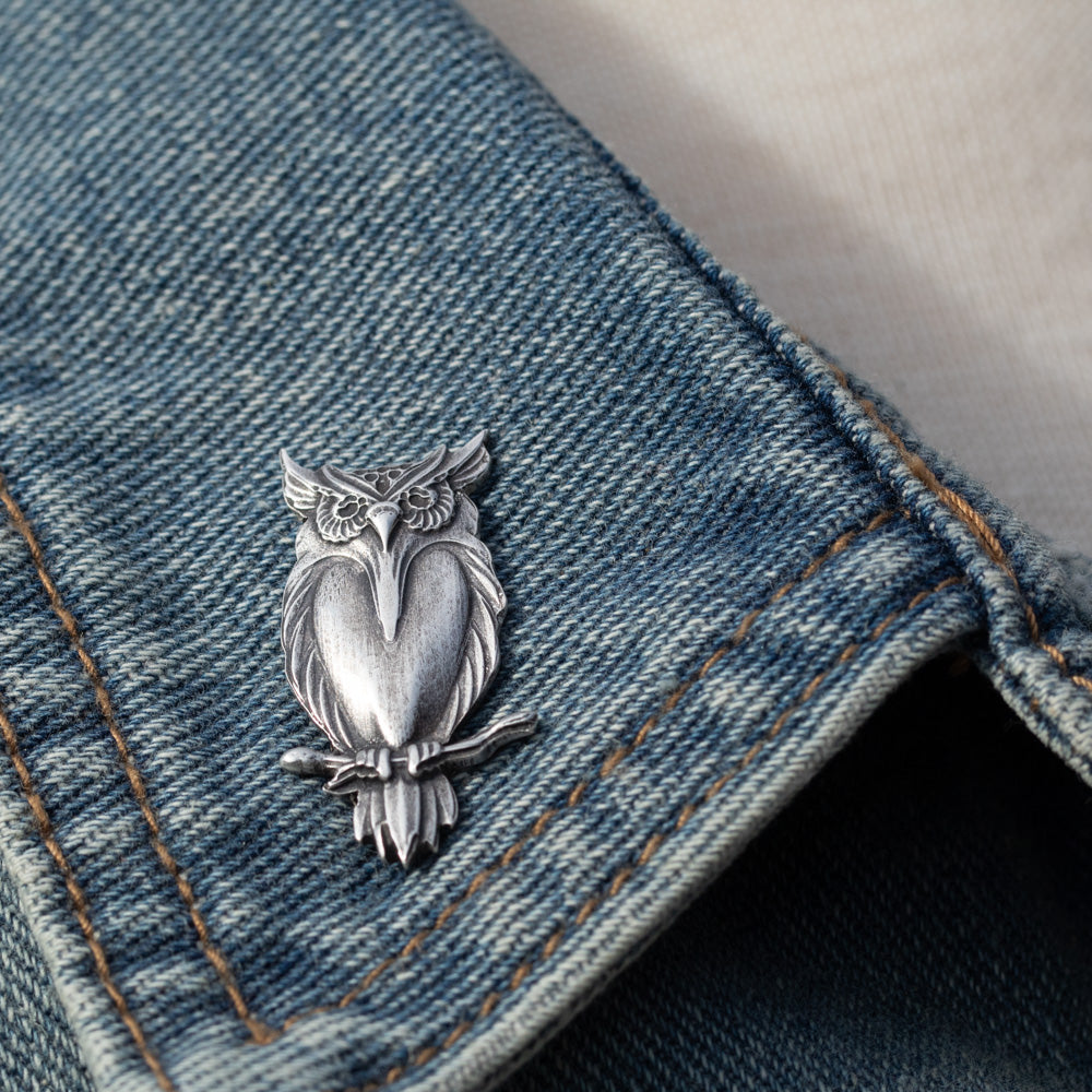 A silver owl-shaped lapel pin resting on a blue denim jacket, with a light-colored shirt visible underneath.