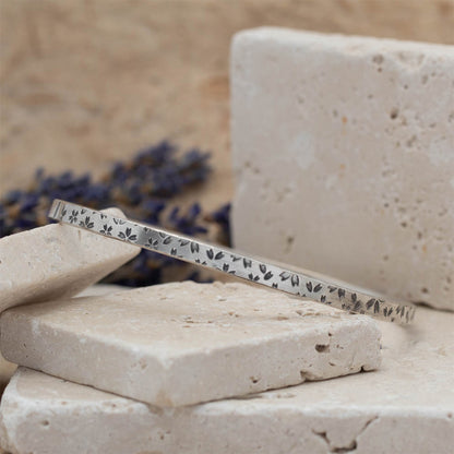 Handmade sterling silver botanical bangle with small floral impressions, photographed on stacked stone blocks with neutral tones and lavender in the background.