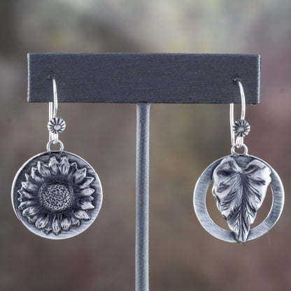 Sterling silver earrings with sunflower and leaf designs on round backings, featuring intricate hand-carved textures, displayed on an earrings stand.