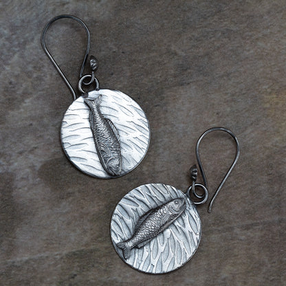 Handcrafted sterling silver earrings featuring detailed fish motifs swimming over textured water-like backgrounds – Swimmers artisan earrings by Divella Designs, displayed on earthy stone surface.