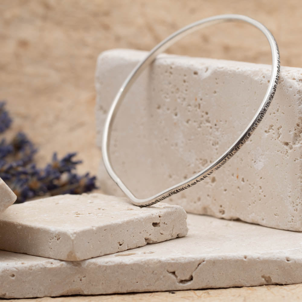 Handmade sterling silver textured bangle photographed on stacked stone blocks with soft neutral styling.