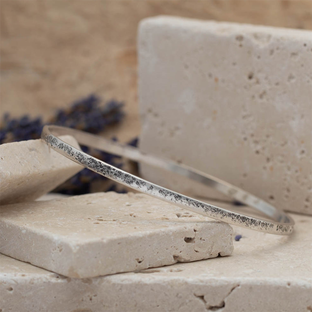 Handmade sterling silver bangle with weathered organic texture, photographed on stacked stone blocks with neutral tones and lavender in the background.
