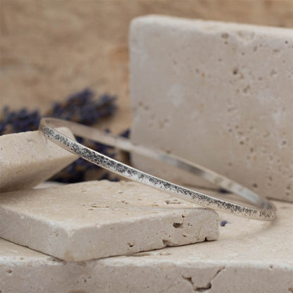 Handmade sterling silver bangle with weathered organic texture, photographed on stacked stone blocks with neutral tones and lavender in the background.