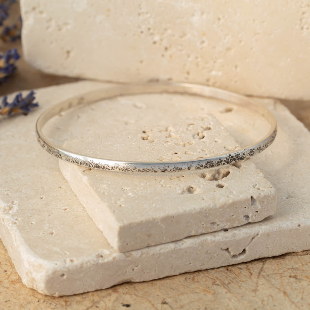 Handmade sterling silver bangle with weathered organic texture, photographed on stacked stone blocks with neutral tones and lavender in the background.