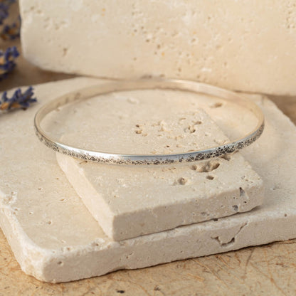 Handmade sterling silver bangle with weathered organic texture, photographed on stacked stone blocks with neutral tones and lavender in the background.