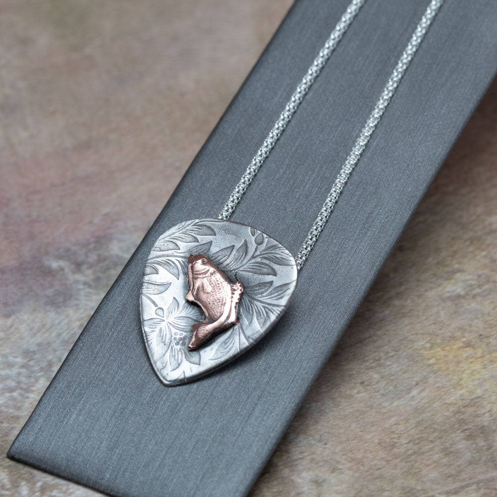 Handcrafted sterling silver pendant with etched floral background and raised copper jumping bass fish, hung on a silver chain – rustic nature-inspired necklace by Divella Designs, displayed on dark backgroun