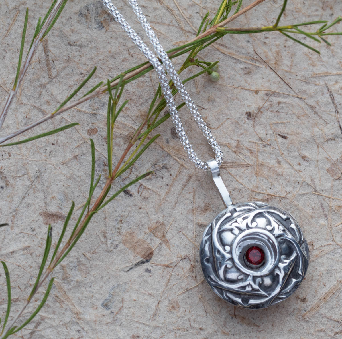 Handcrafted sterling silver pendant necklace featuring a deep red garnet gemstone in a floral shadowbox design, with intricate botanical detailing and oxidized vintage finish