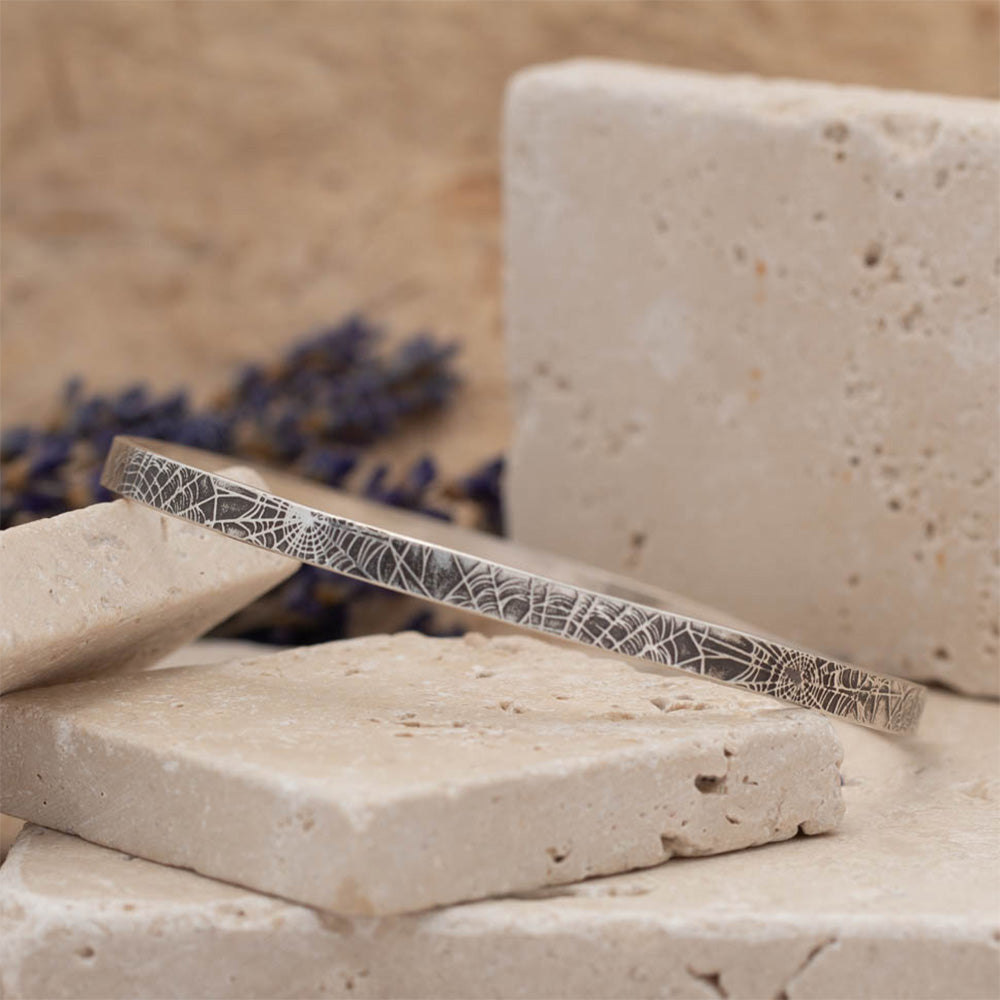 Handmade sterling silver spiderweb textured bangle resting on stacked stone blocks with soft neutral tones and lavender in the background.
