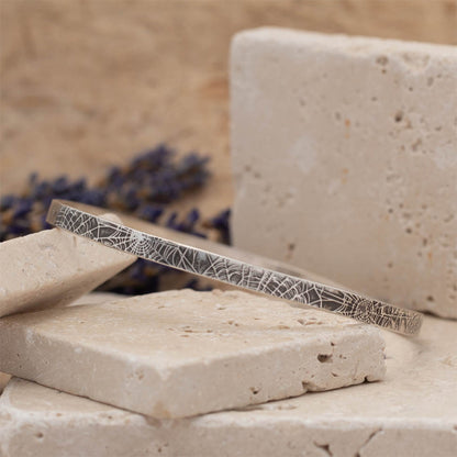 Handmade sterling silver spiderweb textured bangle resting on stacked stone blocks with soft neutral tones and lavender in the background.