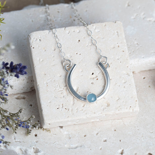 Silver horseshoe necklace with an aquamarine stone on a white platform with lavender flowers.
