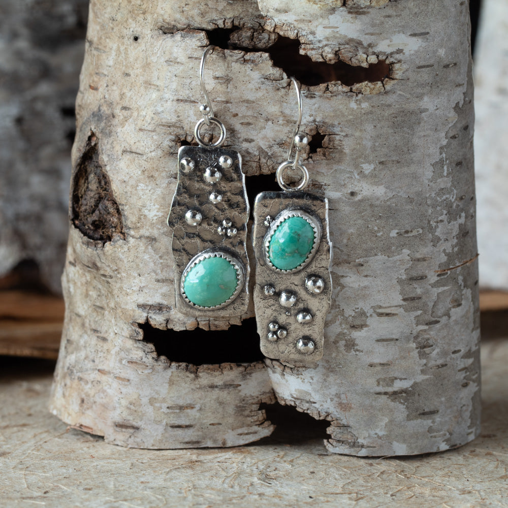 Silver earrings with turquoise stones hanging on a piece of birch wood.