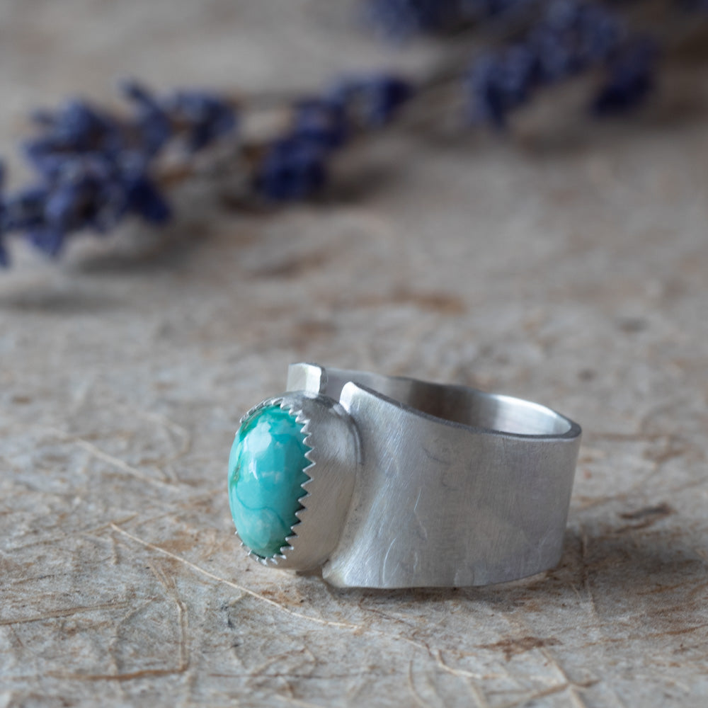 Silver ring with a turquoise stone on a textured surface with blurred flowers in the background