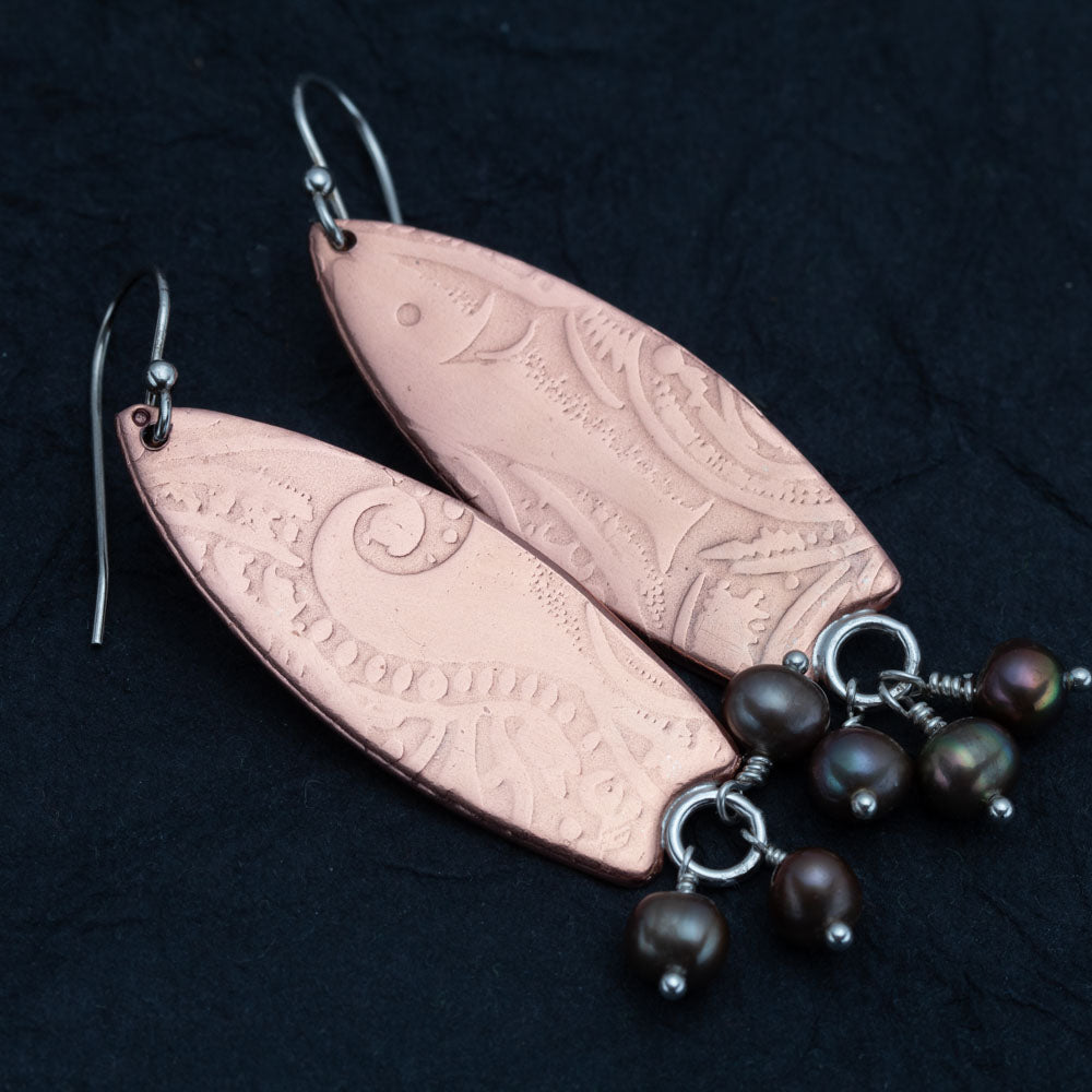 "Handcrafted copper earrings with surfboard shape and embossed pattern, accented with dangling dark freshwater pearls – Surf’s Up artisan design by Divella Designs, shown on black background.