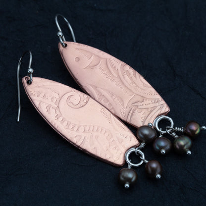 "Handcrafted copper earrings with surfboard shape and embossed pattern, accented with dangling dark freshwater pearls – Surf’s Up artisan design by Divella Designs, shown on black background.