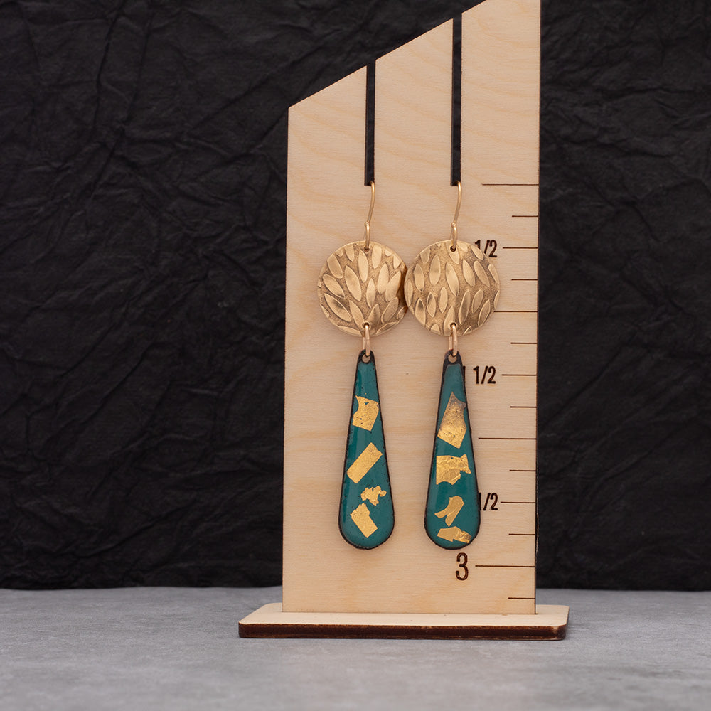 Teal and gold enamel teardrop earrings with round textured brass accents displayed on a gray jewelry stand.