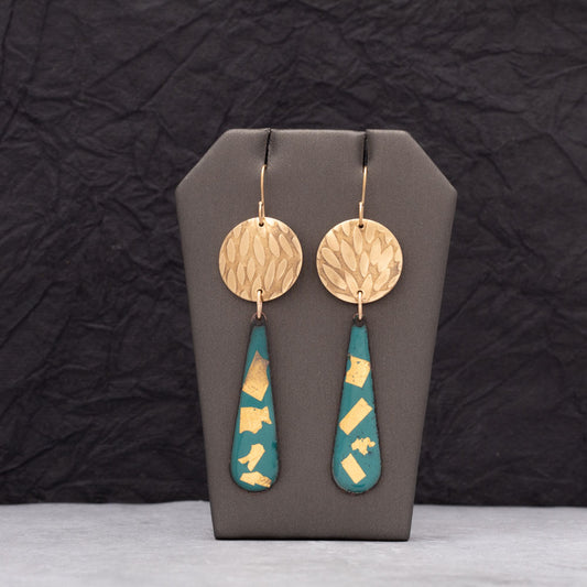 Teal enamel teardrop earrings adorned with 24k gold flakes and paired with round textured brass discs, displayed on a gray jewelry stand against a dark crinkled background.