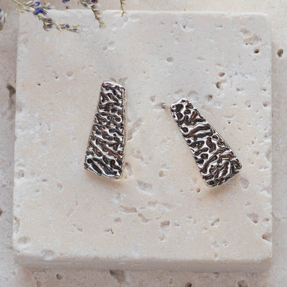 Close up of handmade fine silver stud earrings with textured tree bark pattern, featuring trapezoid shape and rustic organic design for nature-inspired artisan style.