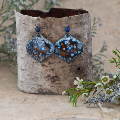 Tidal Pool Enamel Earrings – handcrafted diamond-shaped copper earrings with ocean blue, white, and rust-toned enamel glaze, accented by blue gemstone beads. Displayed on rustic birch bark with wildflowers.