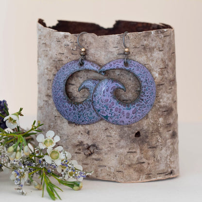 "Handcrafted enamel earrings with a swirling wave design in soft lavender and indigo tones, featuring floral patterns, displayed on birch bark with delicate white wildflowers – Tideflower Spiral by Divella Designs.