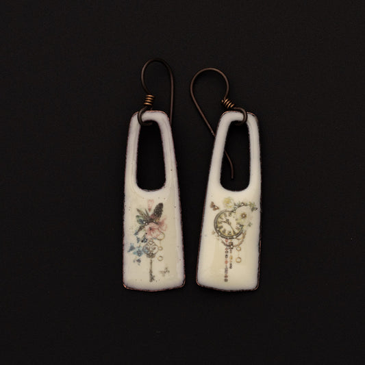 Rectangular white enamel earrings with open oval tops, featuring delicate printed designs of a dragonfly and crescent moon with botanical and celestial motifs, displayed on a black background.
