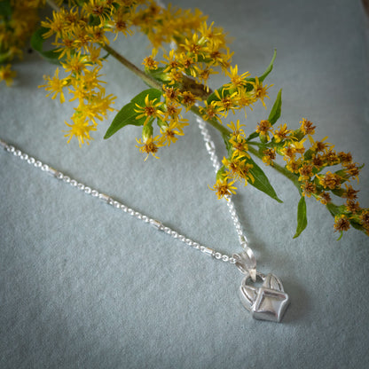 A sterling silver chain necklace with a small geometric fox face pendant, styled on a textured green surface with yellow wildflowers.