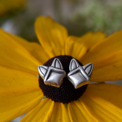 A pair of sterling silver geometric stud earrings shaped like stylized fox heads, displayed on a dark flower center with bright yellow petals in the background.