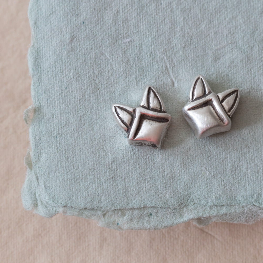 A pair of small sterling silver stud earrings shaped like stylized fox heads, resting on a pale blue textured fabric surface against a soft beige background.